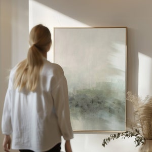May include: A person stands in front of a framed abstract painting with a light green and gray color scheme. The painting is hanging on a white wall.