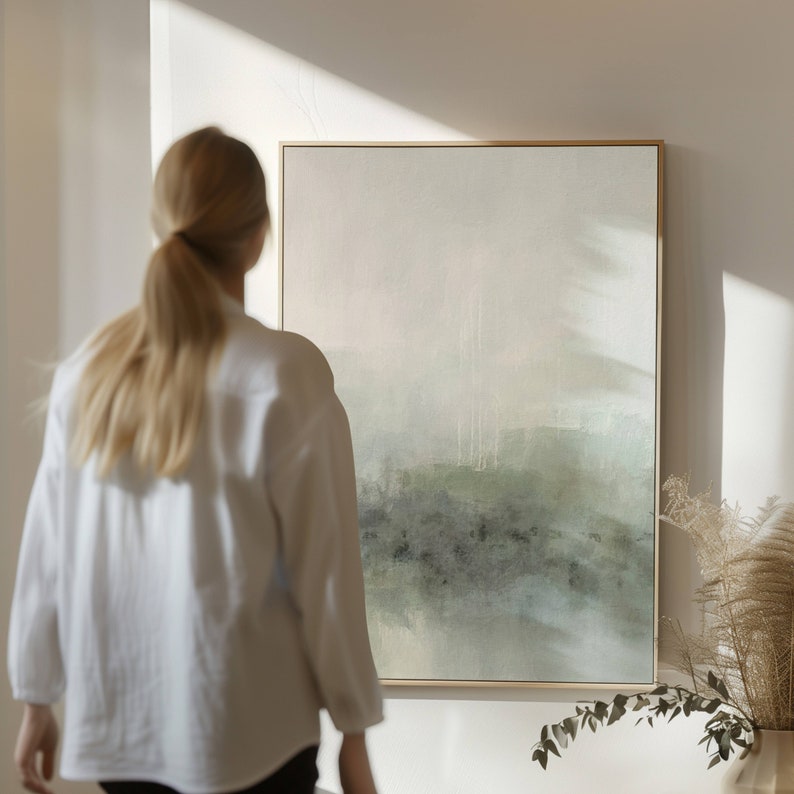 May include: A person stands in front of a framed abstract painting with a light green and gray color scheme. The painting is hanging on a white wall.