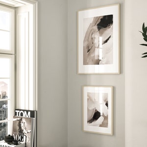 May include: Two framed abstract art prints in a neutral color palette of beige, brown, and black. The prints are hung on a white wall.