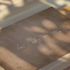 May include: A brown paper card with the handwritten text "how lucky are we" in white cursive. The card is angled, with shadows from overhead creating a natural, soft aesthetic.