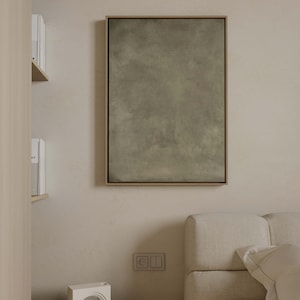 May include: A framed abstract art piece with a muted green and gray color palette. The artwork is rectangular and hangs on a beige wall. The frame is a light brown color. The image is in a bedroom setting.