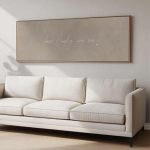 May include: A rectangular framed artwork with the words "how lucky are we" in white script, and a small heart. The artwork has a beige background and a light brown frame. Below, a white sofa with three cushions.