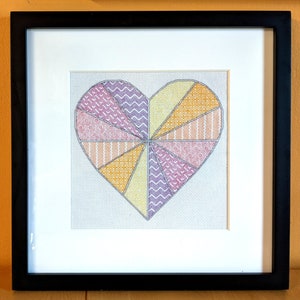 May include: Framed cross-stitch heart artwork. The heart is divided into sections with various patterns and colors, including orange, yellow, and purple. The artwork is framed in a black frame with a white mat.