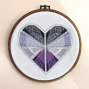 May include: A heart-shaped cross-stitch embroidery in a wooden hoop. The heart is made up of geometric shapes in shades of gray, purple, and white.