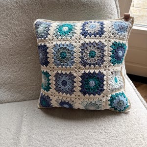 Op de afbeelding: Vierkant gehaakt kussen met een bloemenpatroon in blauw- en groentinten op een gebroken witte achtergrond. Het kussen ligt op een lichtgrijze gestoffeerde ondergrond. Een klein leren label is aan de hoek bevestigd.