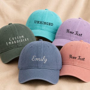 Peut inclure: Cinq casquettes de baseball de différentes couleurs, notamment vert foncé, bleu sarcelle, lavande, bleu et corail. Chaque casquette présente un texte brodé sur le devant : CUSTOM EMBROIDERY, UNHINGED, Your Text, Emily et Your Text.