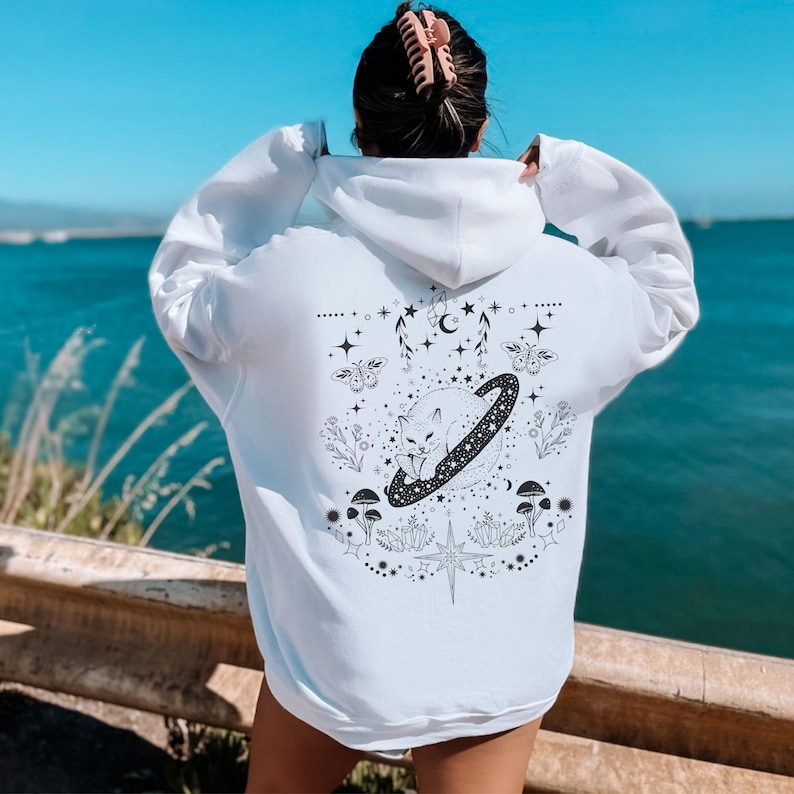 May include: White hoodie with a black and white graphic on the back. The graphic features a fox inside a ringed planet, surrounded by stars, mushrooms, and floral elements. The hoodie has a drawstring hood and long sleeves.
