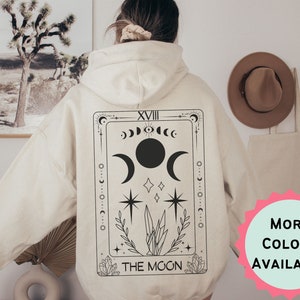 Peut inclure: Un sweat à capuche crème avec un motif de carte de tarot noir et blanc au dos. La carte est la carte "La Lune" et représente un croissant de lune, une pleine lune et une étoile. Le texte "XVIII LA LUNE" est imprimé en bas de la carte.