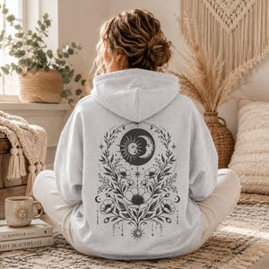 Może przedstawiać: Jasnoszara bluza z kapturem z dużą grafiką w stylu czarnego tuszu na plecach. Wz&oacute;r przedstawia słońce i p&oacute;łksiężyc u g&oacute;ry, otoczone misternymi kwiatowymi pnączami, liśćmi i małymi gwiazdkami w symetrycznym układzie na całej powierzchni.