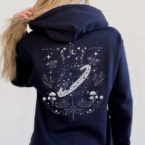 May include: Navy blue hoodie with a white graphic of a cat sleeping on a planet with a ring, surrounded by stars, butterflies, and celestial elements. The design is centered on the back of the hoodie.