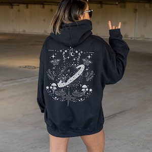 May include: Black hoodie with a white graphic on the back featuring a cat inside a ringed planet, surrounded by stars, moons, butterflies, and mushrooms. The hoodie has a hood and long sleeves. The person in the image is wearing sunglasses.