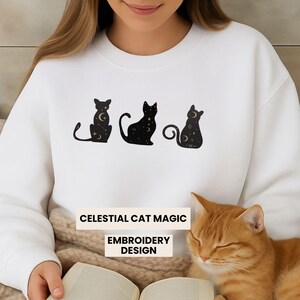 May include: White sweatshirt featuring three black cat silhouettes with celestial moon and star designs. The text "CELESTIAL CAT MAGIC" and "EMBROIDERY DESIGN" are printed below the cats.