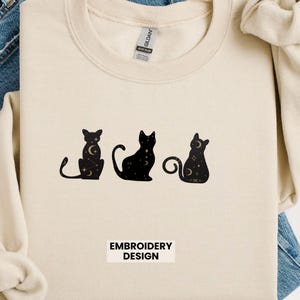 May include: Cream-colored sweatshirt featuring three black cat silhouettes with celestial designs. The cats have moon and star patterns. The text "EMBROIDERY DESIGN" is printed below the cats.