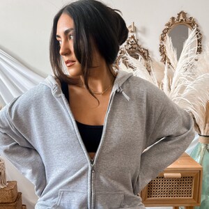 May include: A light gray zip-up hoodie with a drawstring hood and front pockets. The hoodie is made of a soft material and has a silver zipper. The person is wearing a black top underneath.