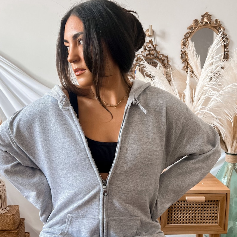 May include: A light gray zip-up hoodie with a drawstring hood and front pockets. The hoodie is made of a soft material and has a silver zipper. The person is wearing a black top underneath.