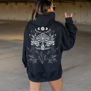 May include: Black hoodie with a white graphic design on the back. The design features a moth, crescent moons, stars, and botanical elements. The hoodie has a hood and a relaxed fit.