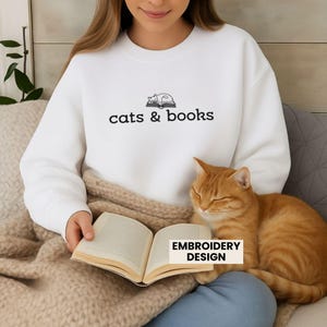 May include: White sweatshirt with the text "cats & books" and a graphic of a cat sleeping on a book. An orange tabby cat is resting on a person's lap, next to an open book. The words "EMBROIDERY DESIGN" are in a white rectangle.