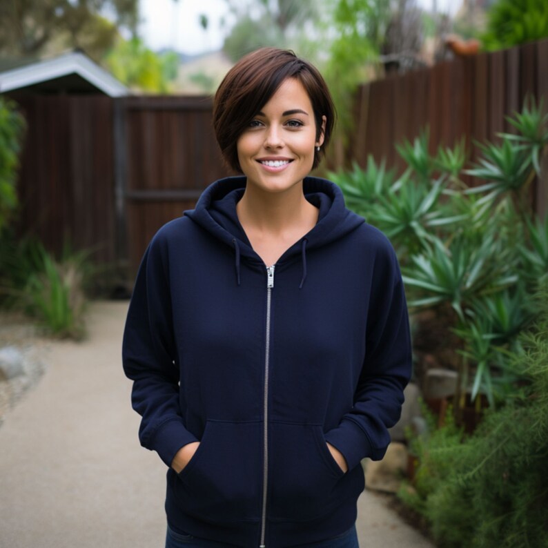 May include: A woman wearing a dark blue zip-up hoodie with a hood. She is standing in front of a wooden fence and green plants.