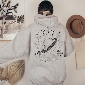 May include: A light gray hoodie with a black graphic design on the back. The design features a cat inside a ringed planet, surrounded by stars, mushrooms, and floral elements. The hoodie has a hood and long sleeves.