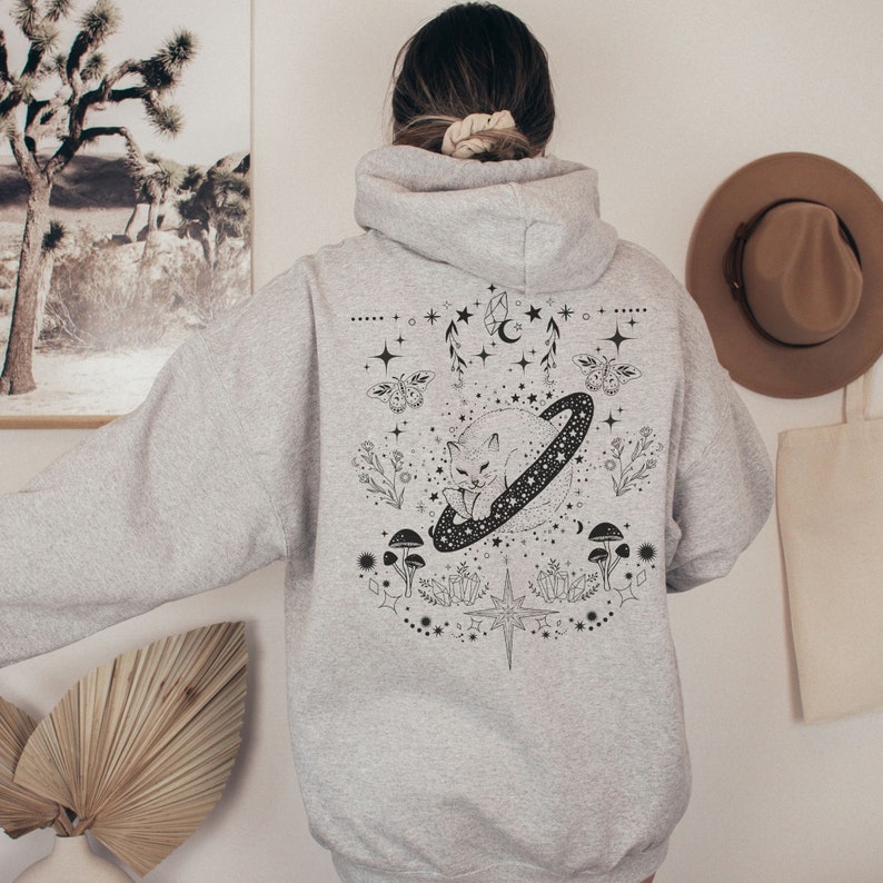 May include: A light gray hoodie with a black graphic design on the back. The design features a cat inside a ringed planet, surrounded by stars, mushrooms, and floral elements. The hoodie has a hood and long sleeves.