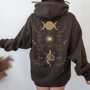 May include: A dark brown hoodie featuring a gold celestial design on the back. The design includes crescent moons, stars, an eye symbol, and a snake. The hoodie has a drawstring hood and a kangaroo pocket.