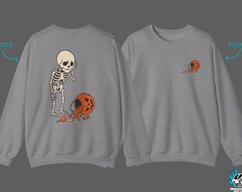 May include: A gray sweatshirt featuring a Halloween-themed design. The back of the sweatshirt displays a sad skeleton with a spilled jack-o'-lantern bucket and scattered candy. The front has a smaller version of the design. A fun, spooky apparel item.