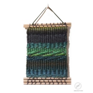 May include: A rectangular woven wall hanging with a gradient of colors, including shades of green, blue, and gray. The woven piece is suspended by two wooden dowels and a green cord. The piece is approximately 10 inches wide and 14 inches long.