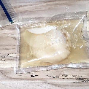 May include: A clear plastic bag with a white, opaque, round object inside. The object is partially submerged in a clear liquid.