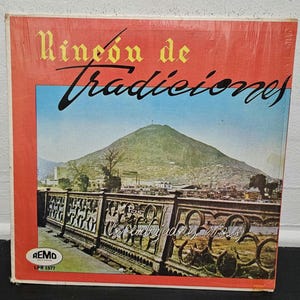 May include: A square vinyl record cover titled Rincón de Tradiciones by Los Embajadores Criollos. The cover features a red border, a landscape photograph of a mountain, and a decorative metal fence in the foreground. The Remo logo is in the corner.
