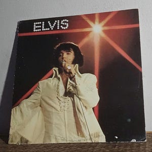 May include: Vintage vinyl record album cover featuring Elvis Presley in a white jumpsuit, singing into a microphone. The album title "ELVIS" is displayed in white block letters at the top. The background is dark with red stage lighting.