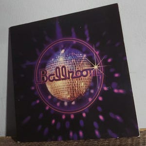 May include: A square, black album cover with a disco ball illustration. The disco ball is in the center, surrounded by a purple circle and the word "Ballroom" in a stylized font. Purple and pink light bursts emanate from the disco ball.