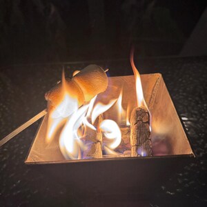 May include: A square metal fire pit with burning wood and flames. Two marshmallows are roasting on a skewer above the flames. The scene is set outdoors at night, with a dark background.