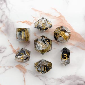 Op de afbeelding: Set van zeven zwarte en gouden dobbelstenen met witte nummers en zilveren vlokken ingebed in de hars. De dobbelstenen zijn in verschillende vormen en maten, waaronder een d4, d6, d8, d10, d12 en d20.