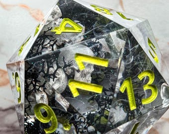 Könnte beinhalten: Ein klarer, vielseitiger Würfel mit neongelben Zahlen. Das Innere zeigt ein schwarz-silbernes Design mit Schädelformen. Der Würfel ist für Tischspiele und Rollenspiele konzipiert.