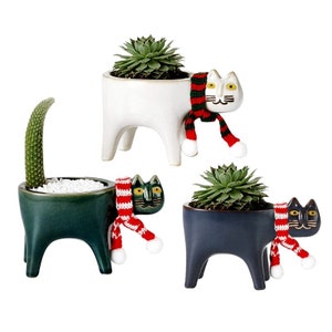 May include: Three ceramic cat planter pots with a red and green scarf. The pots are white, green, and blue. Each pot has a different succulent plant inside.