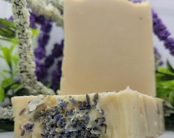 Puede incluir: Dos barras de jabón artesanal, una vertical y otra delante, con capullos de lavanda secos. El jabón es de color beige claro. Flores moradas y blancas en el fondo.