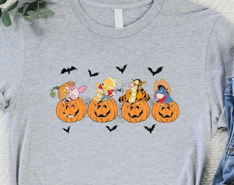 May include: A grey t-shirt featuring a graphic of Winnie the Pooh, Piglet, Tigger, and Eeyore dressed up for Halloween and sitting on pumpkins. The characters are wearing costumes and holding Halloween props.