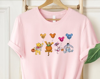 Może przedstawiać: Różowa koszulka z grafiką przedstawiającą Winnie the Pooh, Tigger, Piglet i Eeyore ubranych w kostiumy na Halloween i trzymających balony. Postacie się uśmiechają i mają zamknięte oczy.