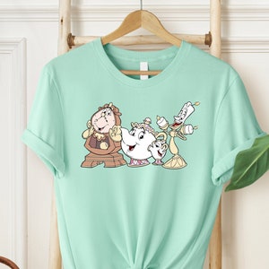May include: A light green t-shirt with a graphic print of characters from the Disney movie Beauty and the Beast. The characters include Cogsworth, Mrs. Potts, and Lumiere.