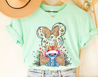 May include: A mint green t-shirt featuring a graphic of Stitch in a Santa hat, inside a Mickey Mouse head outline with a bow. The design includes gingerbread elements and fireworks. A tan fedora hat and a woven purse are also visible.