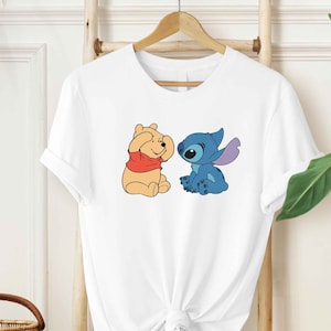 Puede incluir: Camiseta blanca con una imagen de dibujos animados de Winnie the Pooh y Stitch. Winnie the Pooh lleva una camisa roja y Stitch es azul.