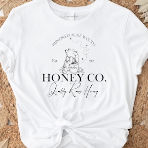 May include: White t-shirt with a black graphic design featuring Winnie the Pooh holding a jar of honey. The graphic includes the text "Hundred Acre Woods", "Est. 1926", "Honey Co.", and "Quality Raw Honey".