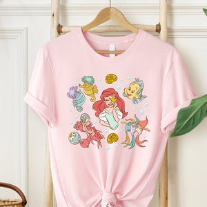 May include: Pink t-shirt with a graphic print of Ariel from The Little Mermaid and other characters from the movie, including Sebastian, Flounder, and a seahorse.