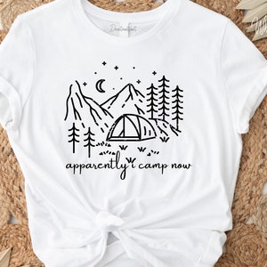 Puede incluir: Camiseta blanca con un dibujo de línea negra de una escena de montaña con una tienda de campaña, árboles y estrellas. El texto "apparently i camp now" está impreso debajo de la imagen.