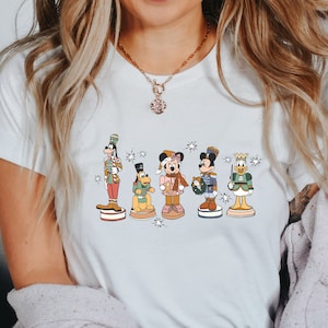 May include: White t-shirt featuring a colourful illustration of Mickey Mouse, Donald Duck, Goofy, Pluto and Minnie Mouse dressed as nutcrackers. The characters are standing in a line and holding musical instruments.