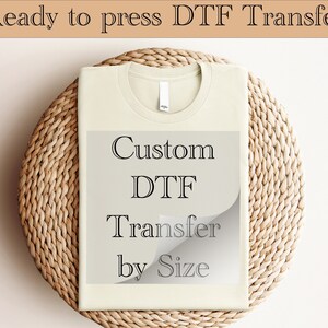 Könnte beinhalten: Ein beiges T-Shirt mit einem weißen Quadrat auf der Vorderseite, auf dem "Custom DTF Transfer by Size" steht. Das T-Shirt liegt auf einer geflochtenen Strohmatte.
