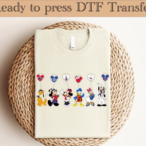 Pode incluir: Uma camiseta de cor creme com um desenho de Mickey Mouse e seus amigos segurando balões. Os balões são vermelhos, brancos e azuis com um design de orelhas de Mickey Mouse. O desenho está centralizado na camiseta.