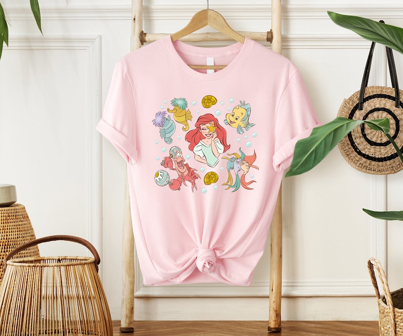 May include: Pink t-shirt with a graphic print of Ariel from The Little Mermaid and other characters from the movie, including Sebastian, Flounder, and a seahorse.