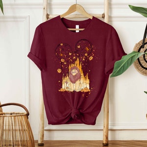 May include: A maroon t-shirt with a graphic print of a lion in front of a castle with fireworks and pumpkins. The castle is a silhouette of a famous theme park.