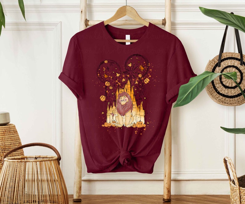 May include: A maroon t-shirt with a graphic print of a lion in front of a castle with fireworks and pumpkins. The castle is a silhouette of a famous theme park.